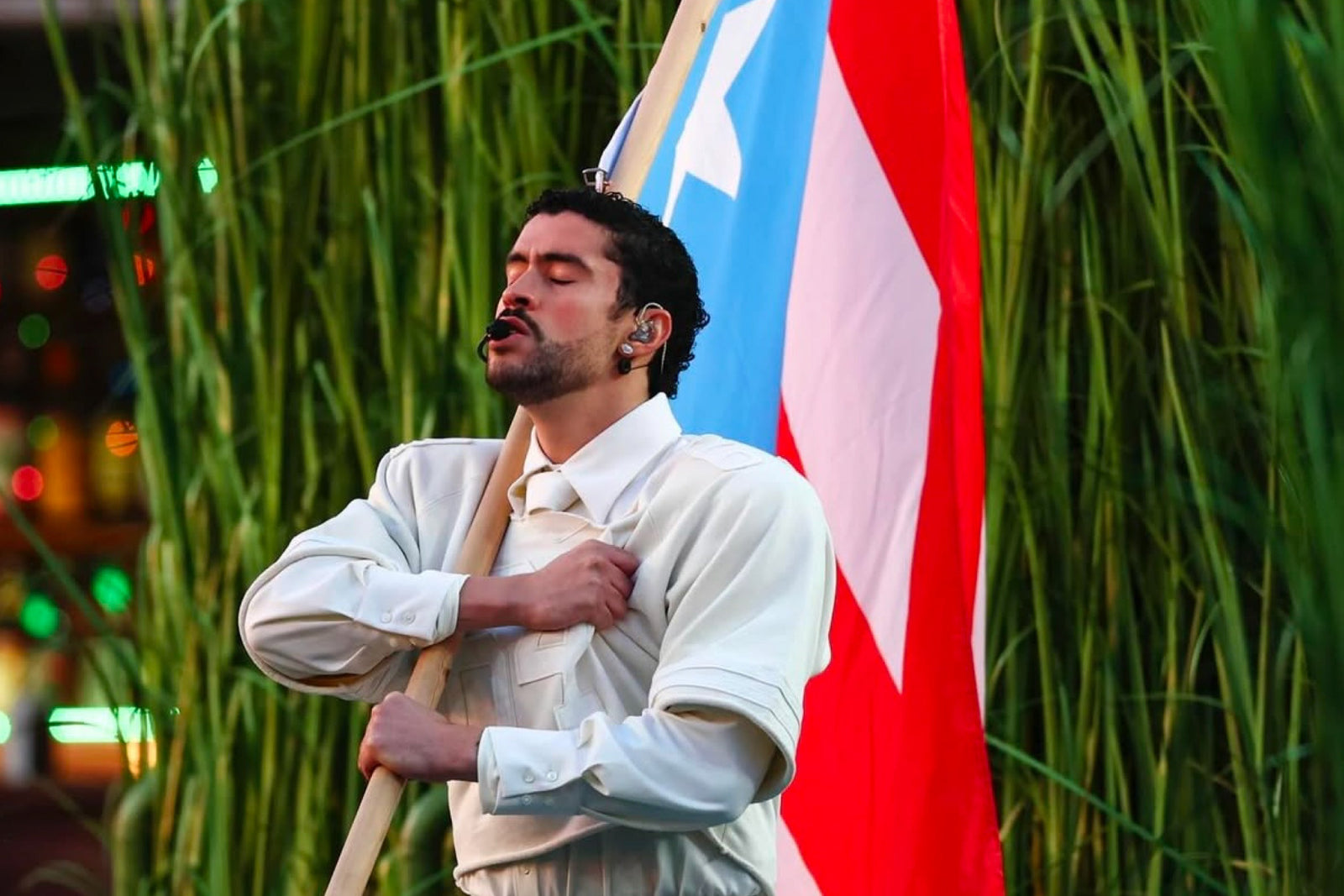 Male performer dressed in white holding a Puerto Rican flag on stage, singing with eyes closed in front of tall green plants.