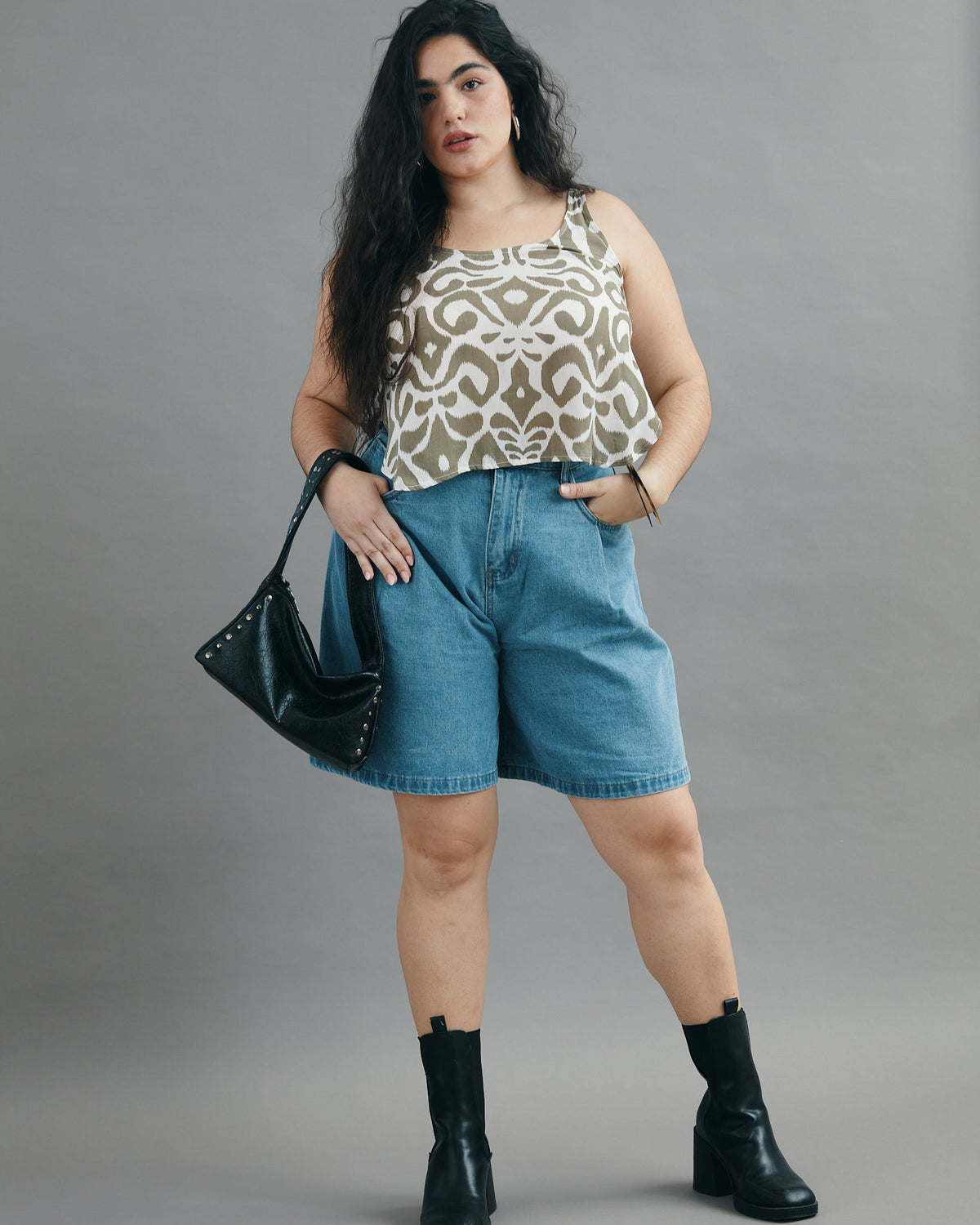 Woman wearing a patterned top, denim shorts, and black boots against a gray background