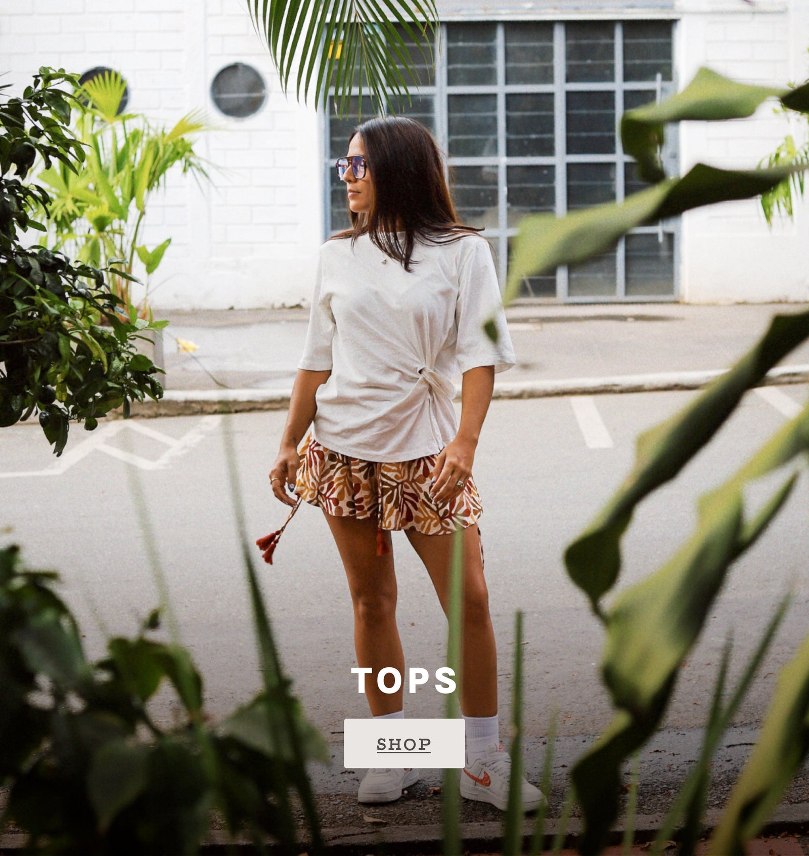 Shop Tops. A woman stands outdoors on a street wearing a white knotted short-sleeve top, brown and beige patterned shorts, white socks, and white sneakers. Large green leaves frame the foreground, with a white building in the background.