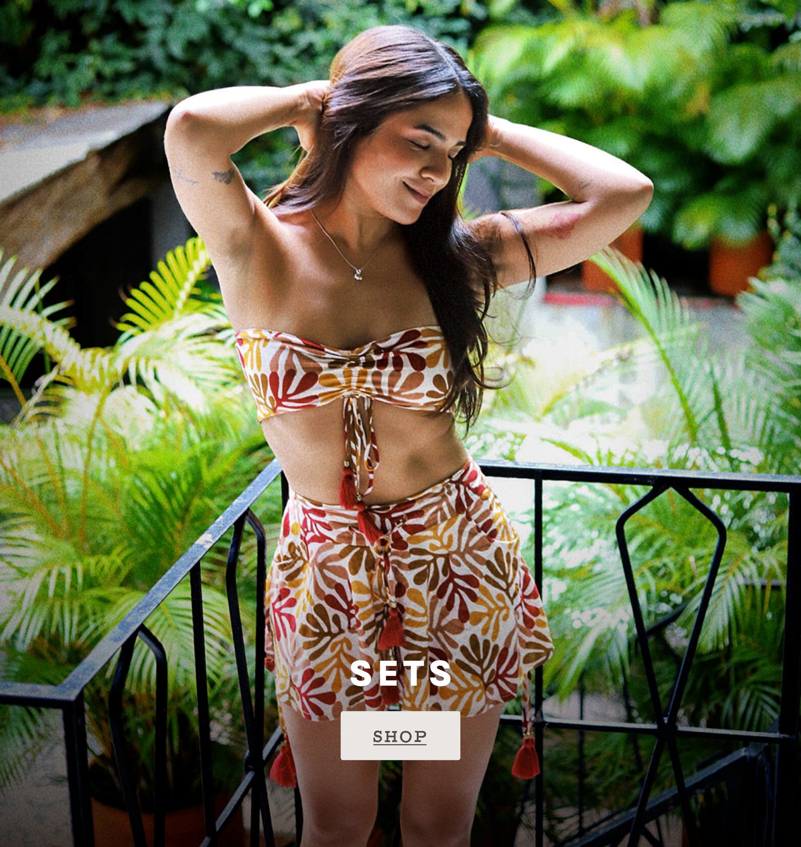 Shop Sets. A woman posing on a balcony wearing a two-piece tropical print set consisting of a strapless bandeau top and matching shorts. The set features a warm brown, orange, and white palm leaf pattern.