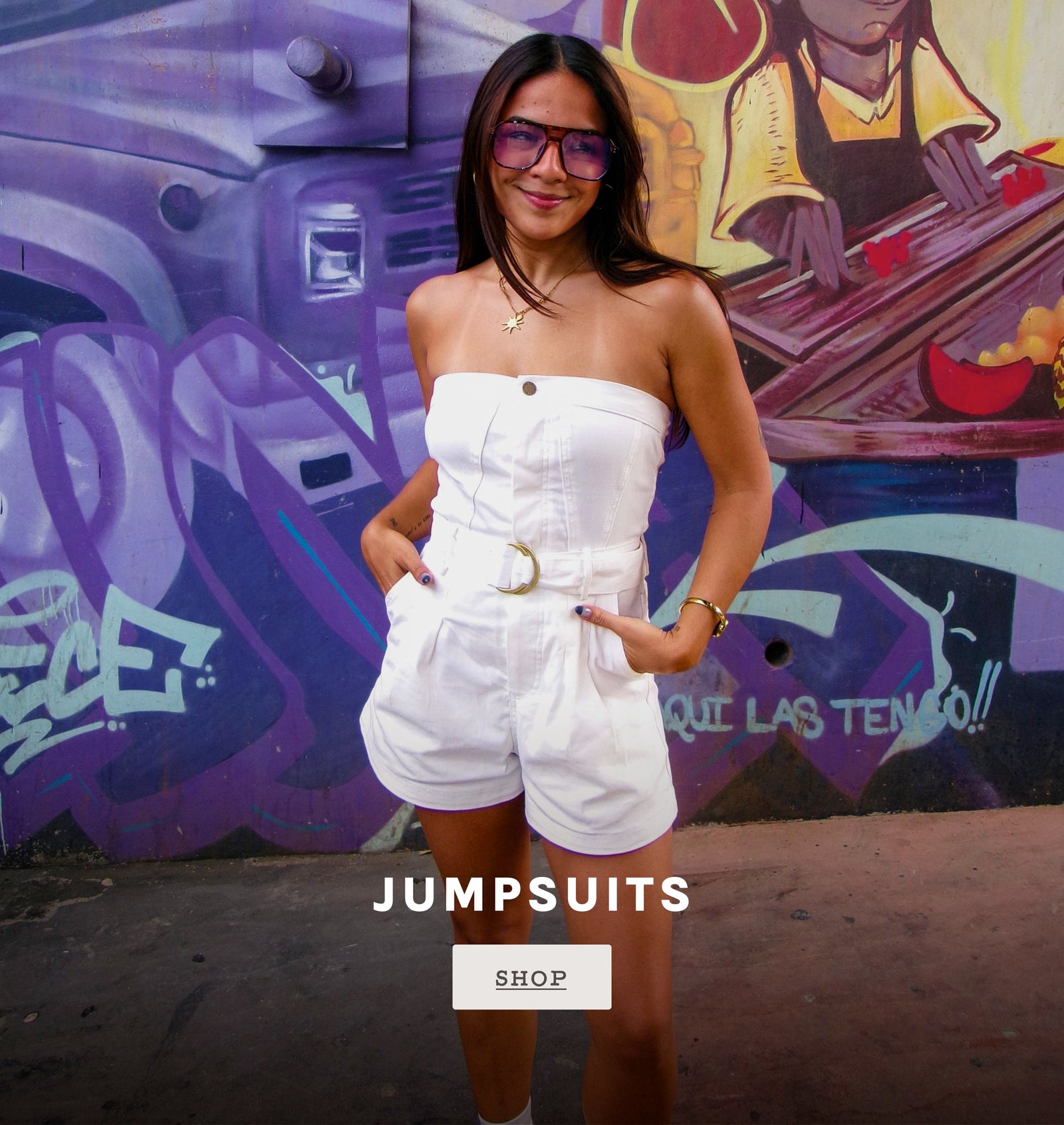 A woman wearing a white strapless jumpsuit with shorts and a gold belt buckle stands in front of a colorful graffiti wall. The image includes a text overlay that reads 'JUMPSUITS' with a 'SHOP' button below.