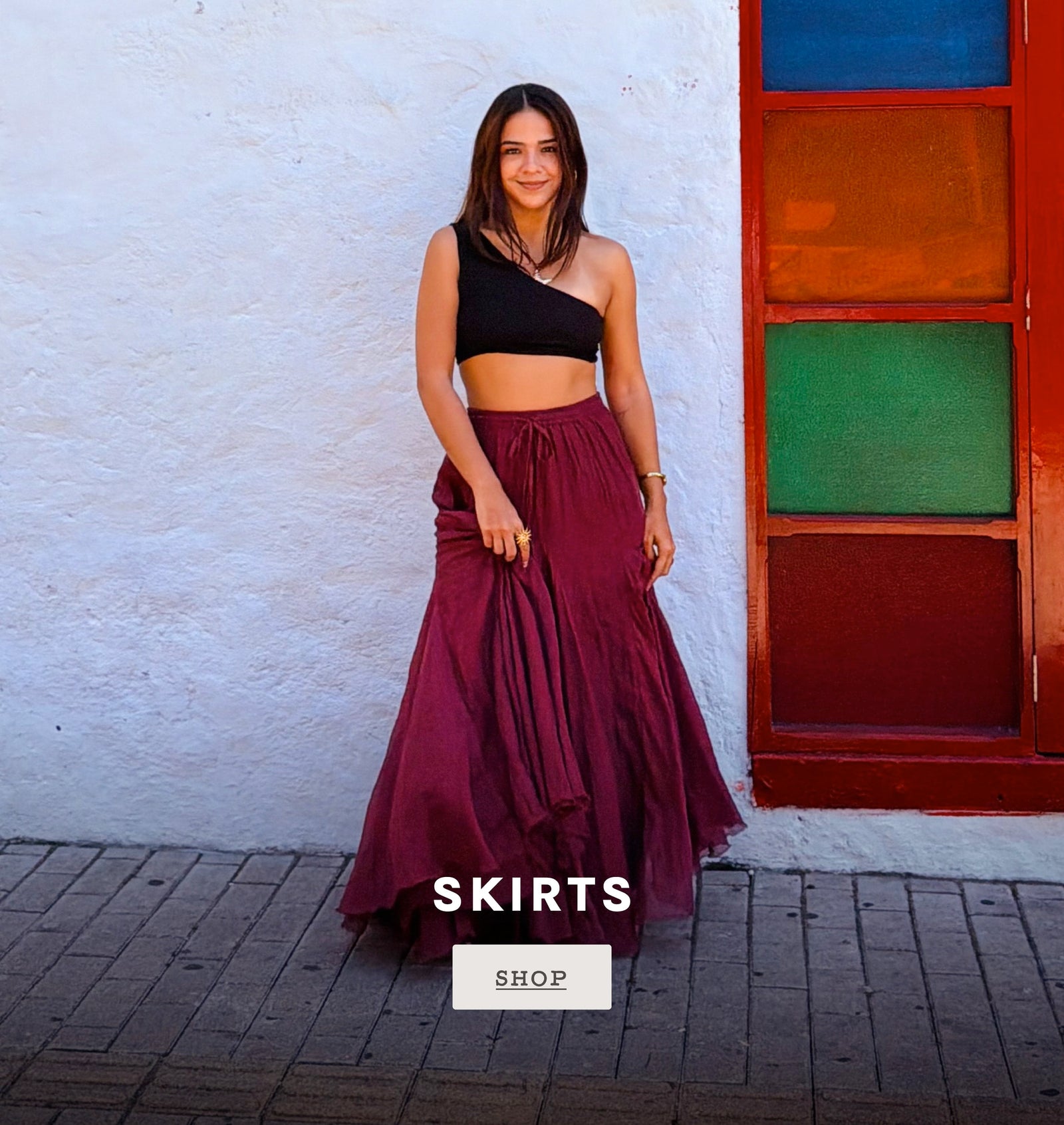 A woman stands outdoors against a white textured wall and a colorful red door, wearing a long flowing dark burgundy skirt with a black one-shoulder top. The image includes the text 'SKIRTS' and a 'SHOP' button overlay.