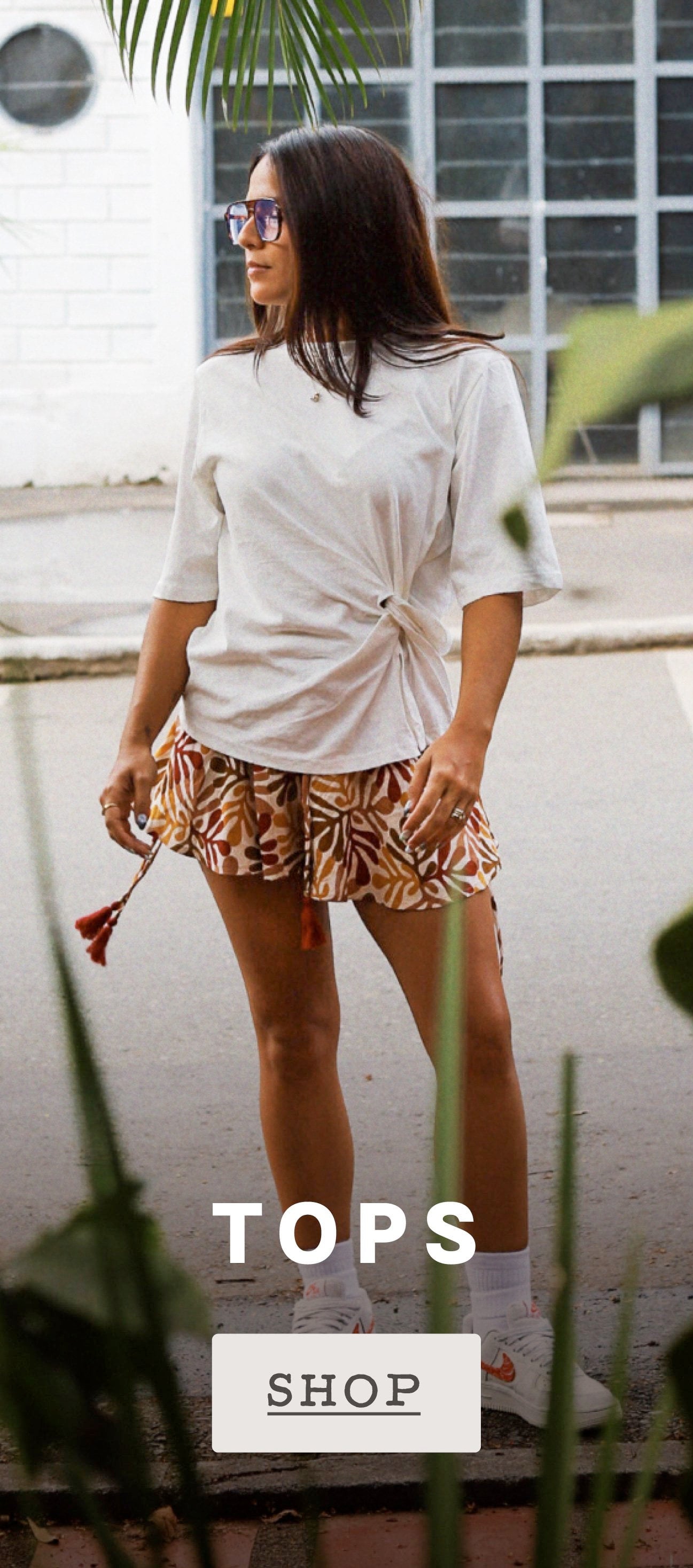 A woman wearing a white oversized t-shirt with a side twist and patterned brown and beige shorts, standing outdoors in front of a building with a window. Text overlays at the bottom read 'SETS' and 'SHOP'.