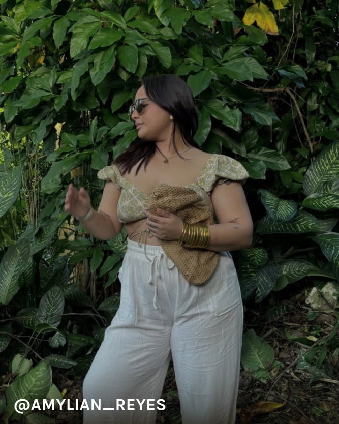 Woman in a gold top and white pants standing in front of green foliage