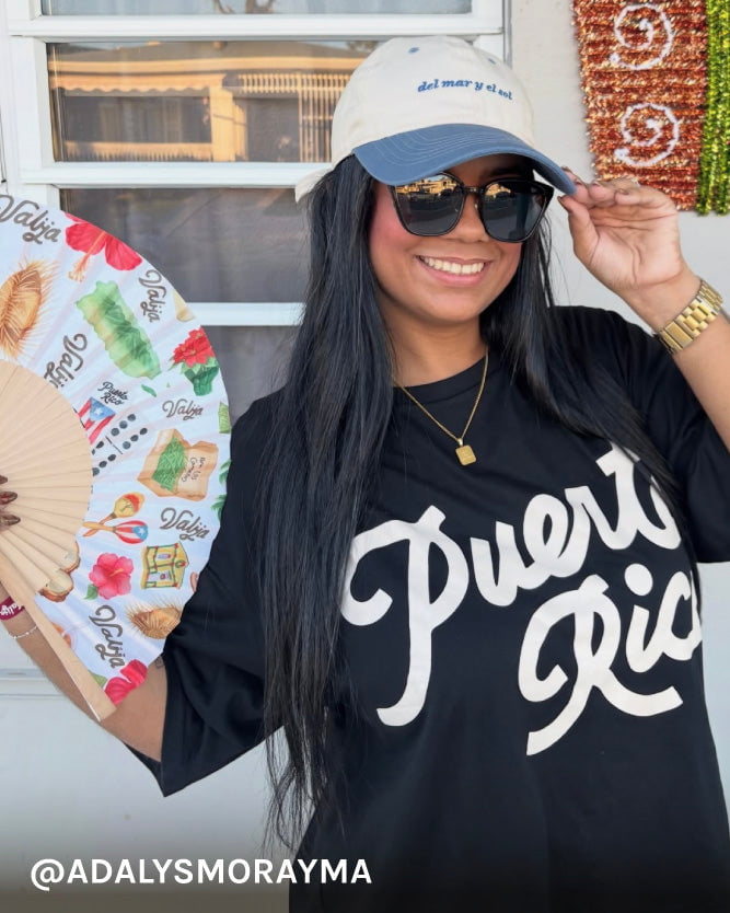 Woman wearing a 'Puerto Rico' shirt and a cap, holding a fan with local designs.