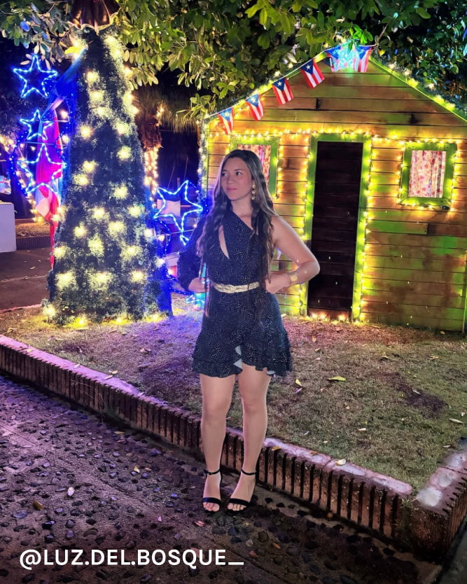 Woman posing in front of a decorated outdoor scene with lights and a small house.